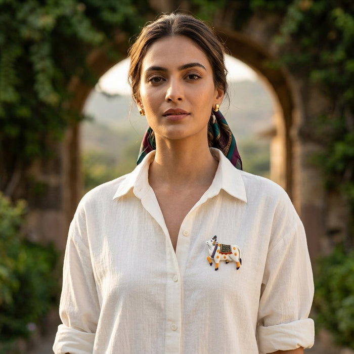 Woman wearing a white shirt with a 925 silver pichwai cow vastra enamel brooch, standing outdoors with greenery in the background